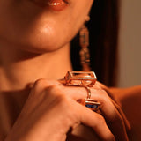 Model holding large gold box ring near chin with drop earrings visible